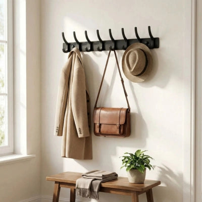 Patère Danielle noire à 8 crochets fixée dans une entrée lumineuse, avec un manteau beige, un sac à main marron et un chapeau suspendus. Décor mur blanc cassé avec un banc en bois et une plante verte.