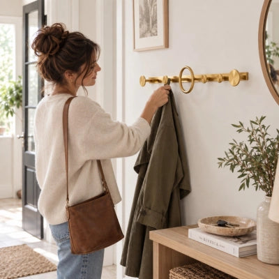 Femme en pull crème accrochant un manteau kaki à la patère murale en laiton Miranda, dans une entrée lumineuse avec console en bois, miroir rond et plante verte.